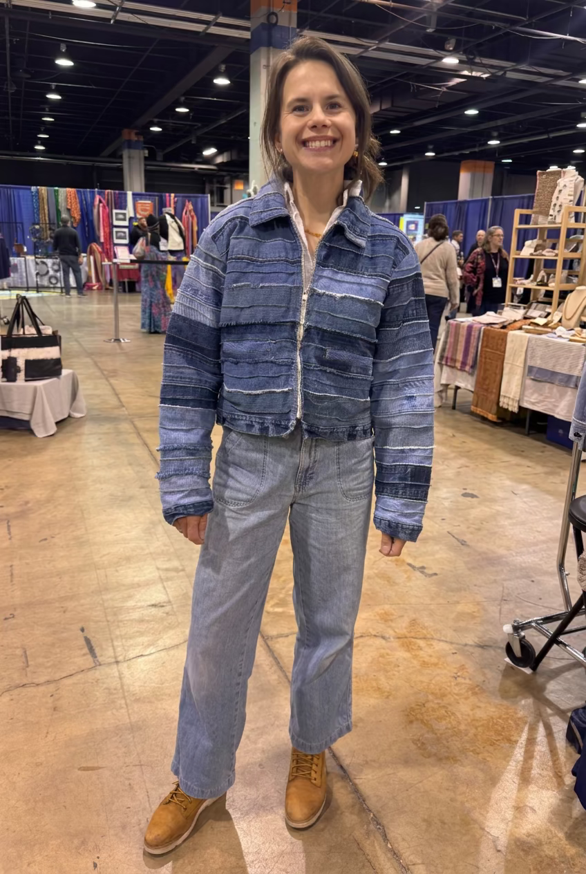 A woman stands in a convention hall wearing a cropped blue denim jacket with horizontal striped patchwork panels, light-wash jeans, and tan lace-up boots. Mannequins and clothing racks are visible in the background.