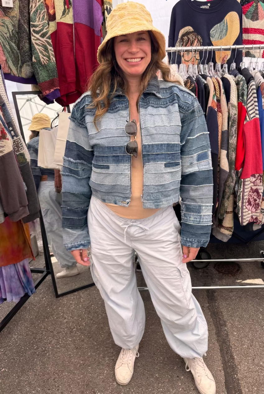 A prenant woman stands at an outdoor market stall wearing a cropped blue striped denim jacket, light gray baggy pants, a yellow bucket hat, and white sneakers. Clothing racks with colorful garments are visible behind her. Cute pregnancy sustainable style